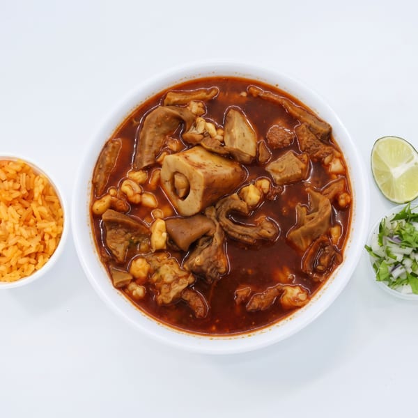 Hearty menudo soup with rice, lime, and chopped onion and cilantro on a white background.