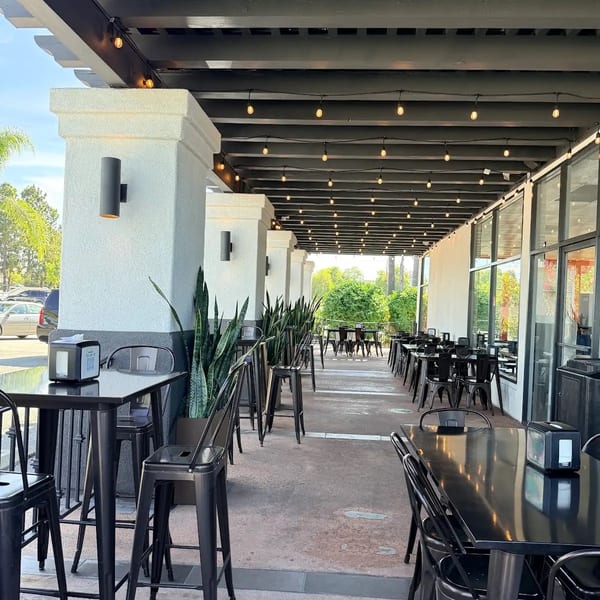 Outdoor patio with black high-top tables, chairs, and string lights under a dark pergola.