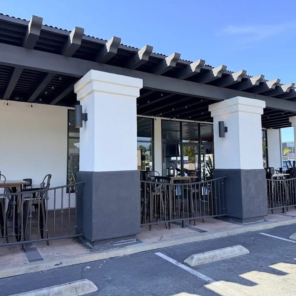Outdoor patio dining area with dark tables and chairs under a pergola, next to a parking lot.