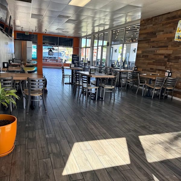 Empty restaurant dining room with dark wood-look floors, tables, chairs, and large windows.