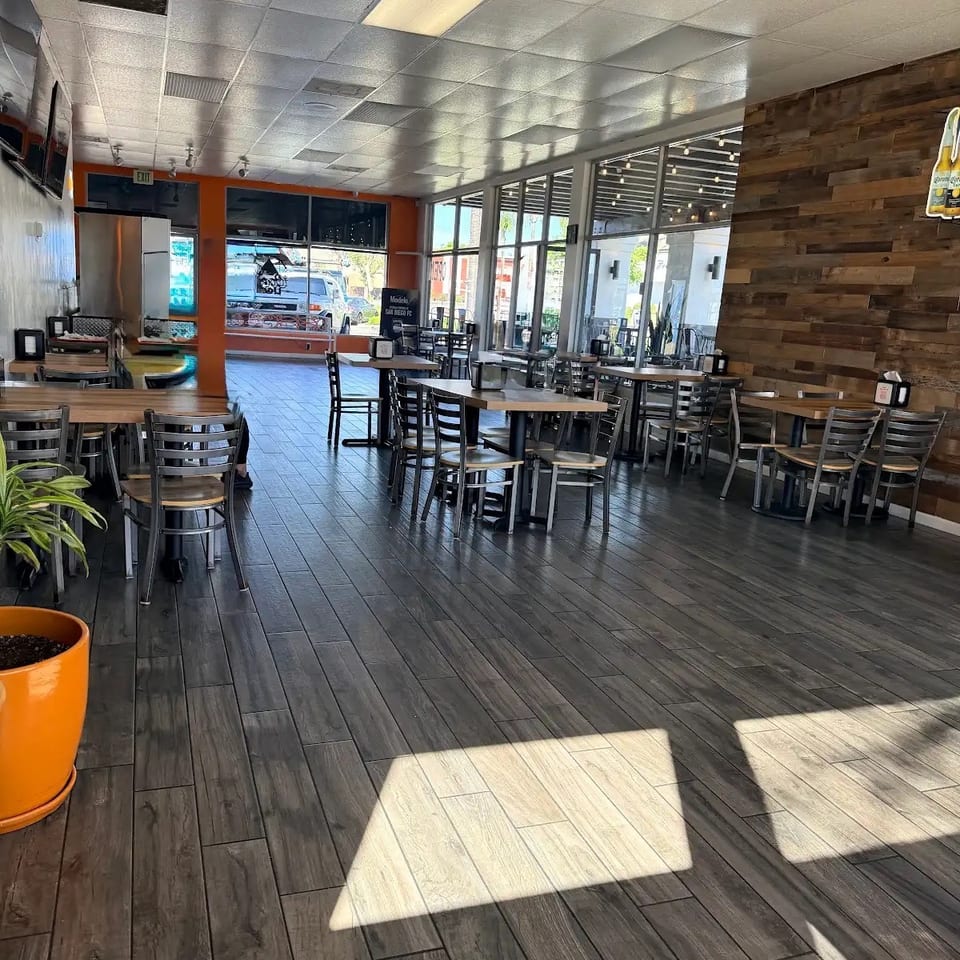 Empty restaurant dining room with dark wood-look floors, tables, chairs, and large windows.