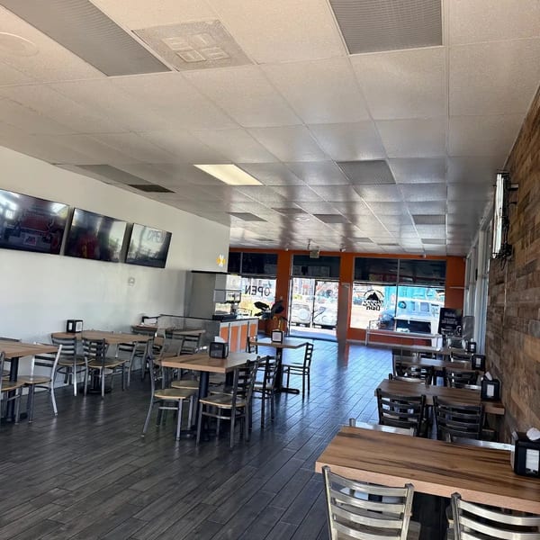 Empty restaurant interior with dark wood-look floors, light walls, and tables with metal chairs.