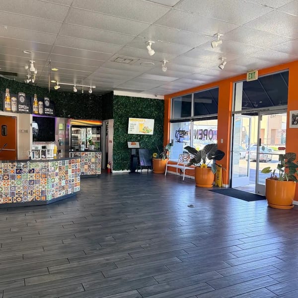 Restaurant interior with a colorful tiled counter, dark wood-look floor, and orange walls with large windows.