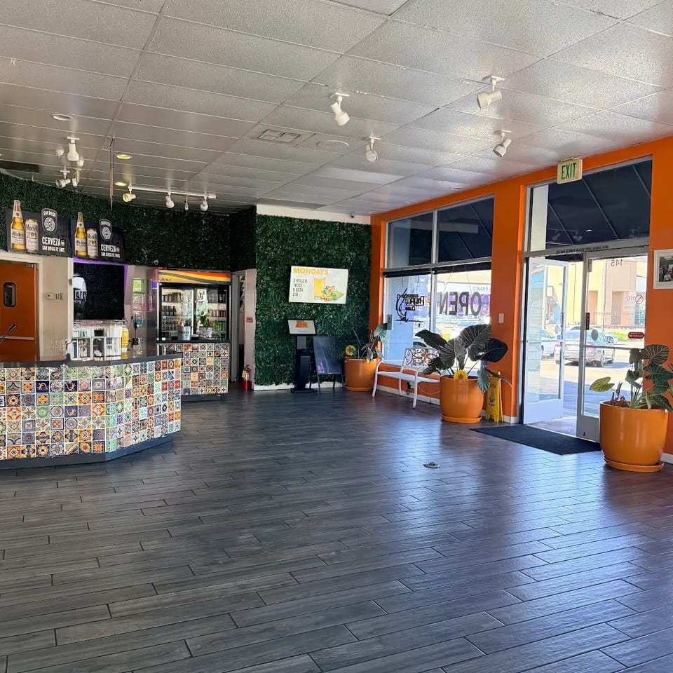 Restaurant interior with a colorful tiled counter, dark wood-look floor, and orange walls with large windows.