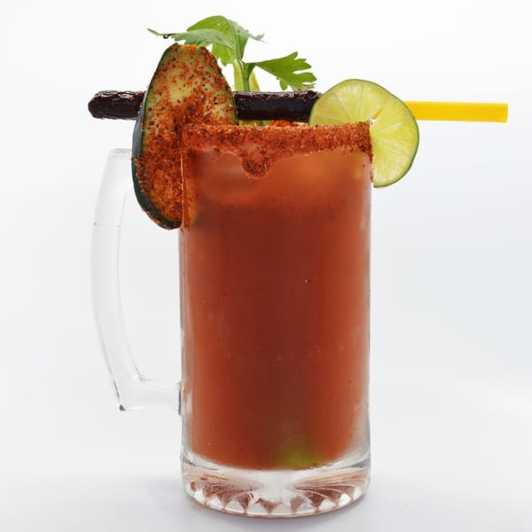 A michelada cocktail in a mug, garnished with chili-dusted cucumber, lime, and a yellow straw.
