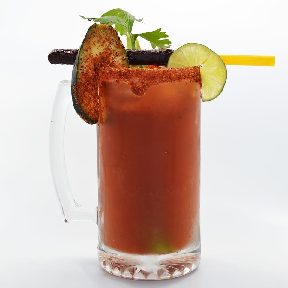 A michelada cocktail in a mug, garnished with chili-dusted cucumber, lime, and a yellow straw.
