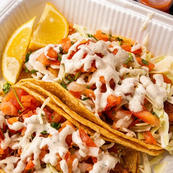 Two fish tacos with cabbage, diced tomatoes, creamy sauce, and orange slices in a takeout container.